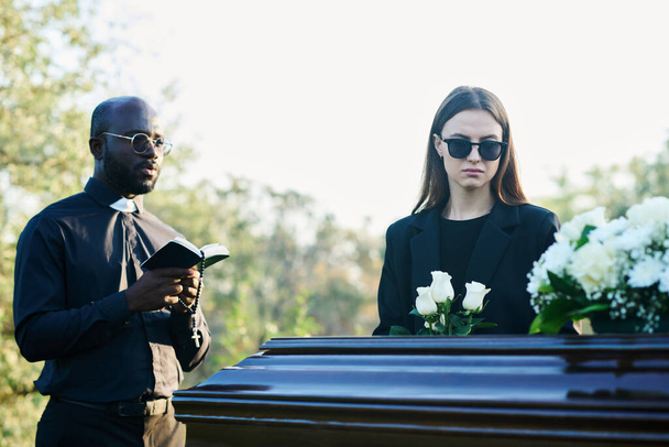Νεαρή απαρηγόρητη χήρα με πένθιμη ενδυμασία και γυαλιά ηλίου που κρατάει ένα μάτσο λευκά τριαντάφυλλα ενώ στέκεται δίπλα στο φέρετρο δίπλα στον ιερέα με τη Βίβλο. - Φωτογραφία, εικόνα