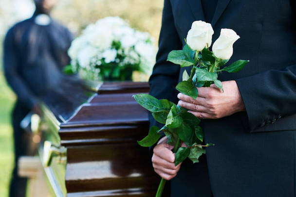 Primer plano del hombre maduro en traje negro sosteniendo dos rosas blancas frescas mientras está de pie frente a la cámara contra el ataúd con tapa cerrada - Foto, Imagen