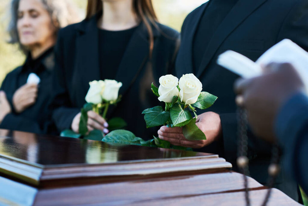 Concentrati su due rose bianche fresche tenute dall'uomo in lutto in abito nero durante il servizio funebre mentre è in piedi vicino alla bara contro sua figlia e sua moglie. - Foto, immagini