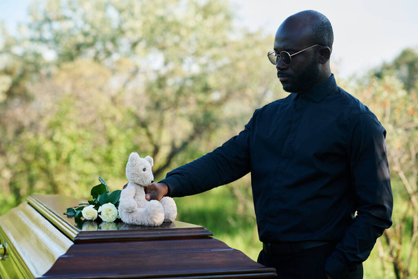 Młody Afroamerykanin w żałobie wkładający białe róże i pluszowego misia na pokrywę trumny na pogrzebie. - Zdjęcie, obraz