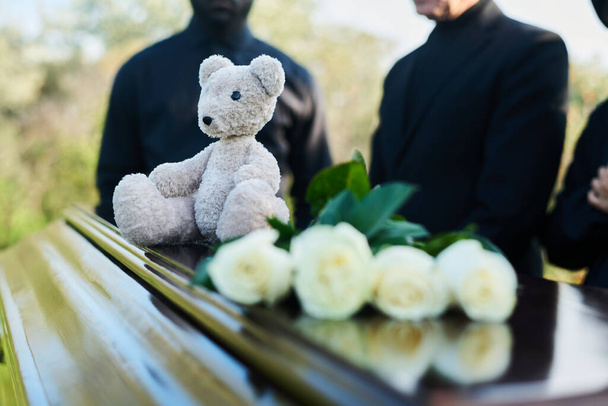 Focus op grijze teddybeer zittend bovenop gesloten doodskist deksel met een bos verse witte rozen die voor de camera staan tegen rouwende mensen - Foto, afbeelding