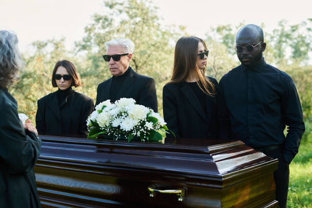 Young woman standing by her grieving husband in mourning clothes during funeral of relative or family member in coffin with white flowers - Photo, Image