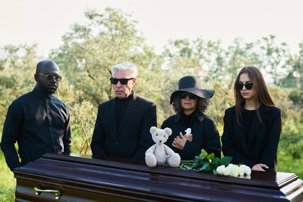 Groupe de personnes interculturelles en deuil dans des vêtements de deuil réunis par cercueil avec ours en peluche et roses blanches fraîches sur le dessus du couvercle fermé - Photo, image