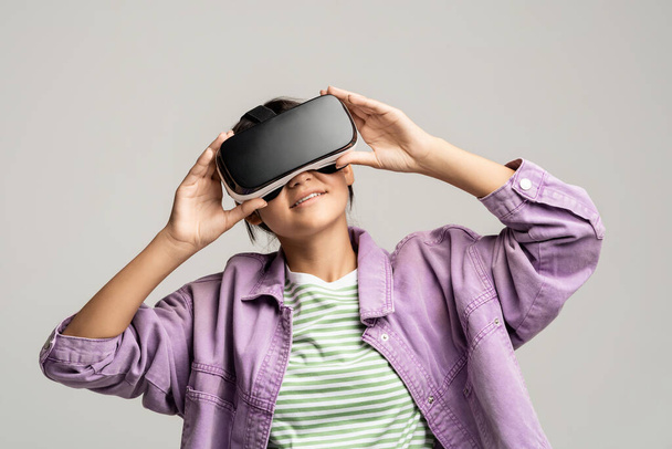 Tiener meisje in virtuele bril bril bril kijken film spelen videospelletjes op grijze achtergrond. Lachende tiener genietend van de virtuele wereld in VR helm. Cyberspace, entertainment, online recreatie concept. - Foto, afbeelding