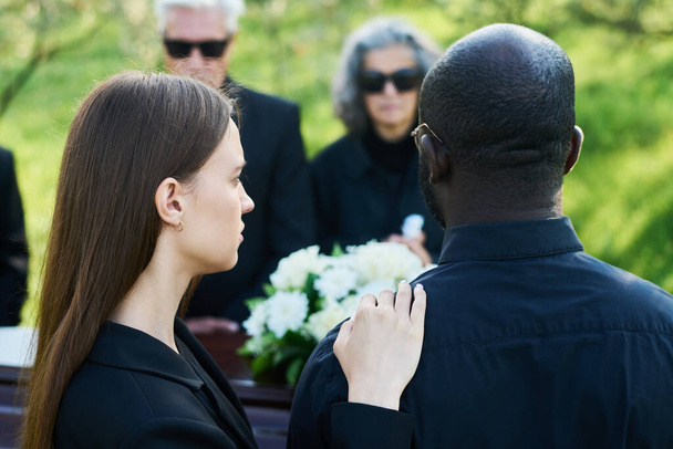 Πίσω όψη της νεαρής γυναίκας που πενθεί κρατώντας το χέρι στον ώμο του συζύγου της με πένθιμη ενδυμασία ενώ και οι δύο στέκονται μπροστά από το φέρετρο - Φωτογραφία, εικόνα
