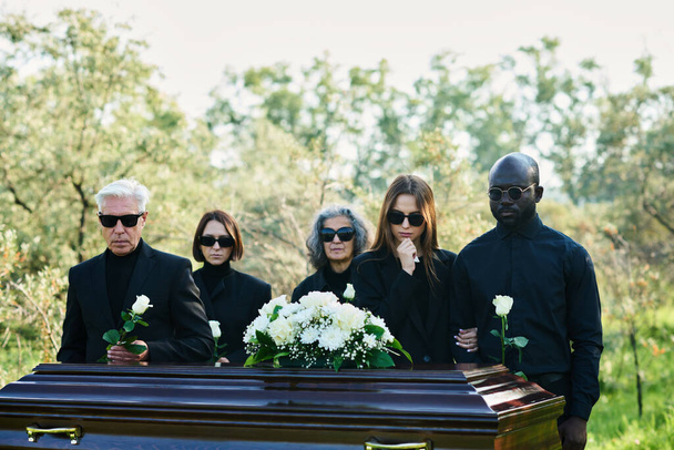 Група міжкультурних засмучених людей у траурному одязі та сонцезахисних окулярах тримає білі троянди, стоячи перед труною з закритою кришкою - Фото, зображення