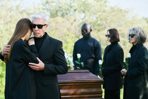 Скорбящий взрослый мужчина в черном костюме обнимает свою скорбящую дочь, выражая свою симпатию к потере дорогого друга или члена семьи - Фото, изображение
