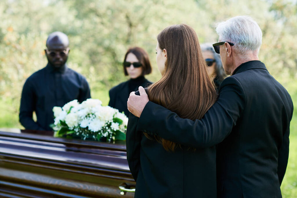 Вид ззаду на зрілого чоловіка в чорному костюмі, який обіймає свою сумну дочку на похоронах і прощання з родичем, другом або членом сім'ї - Фото, зображення
