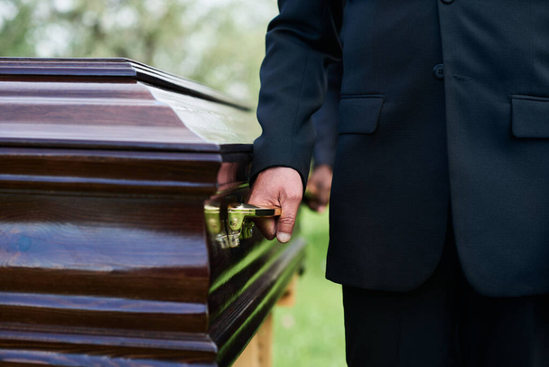 Close-up de homem de terno preto segurando pelo punho de caixão de madeira com pessoa morta enquanto o carrega com outras pessoas na cerimônia fúnebre - Foto, Imagem