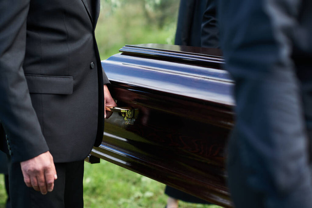 Сосредоточьтесь на человеке в черном костюме, держащем за ручку деревянный гроб, когда несете его вместе с другими людьми после прощальной церемонии. - Фото, изображение
