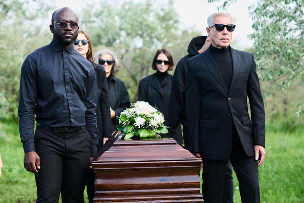 Grupa pogrążonych w żałobie ludzi w żałobie niosących drewnianą trumnę ze świeżymi chryzantemami na wierzchu zamkniętej pokrywy na pogrzebie - Zdjęcie, obraz