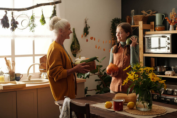Happy grandmother with bowl of fresh vegetables standing in front of cheerful granddaughter holding green zucchini and small pumpkin - Photo, Image