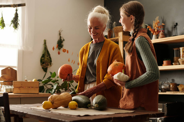Happy aged woman and her cute granddaughter with pigtails having chat while standing by kitchen table and sorting out fresh vegetables - Photo, Image