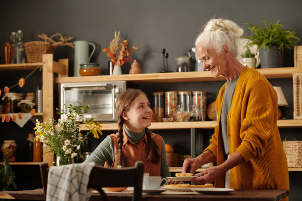 Šťastná roztomilá dívka s copánky při pohledu na babičku, zatímco s čajem s chutným domácím koláčem s jablky pěstovanými v zahradě u chalupy - Fotografie, Obrázek
