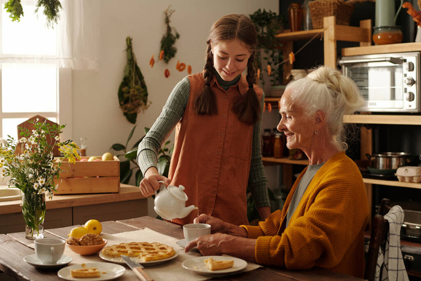 Fille heureuse en tenue décontractée versant une tisane dans la tasse de sa grand-mère assise à côté d'une table en bois servie avec une délicieuse tarte aux pommes maison - Photo, image