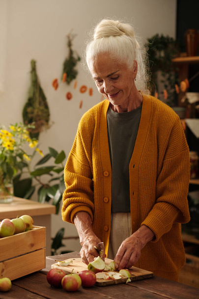 Senior woman with white hair standing by wooden table in front of camera and cutting fresh ripe apples grown in the garden by country house - Fotografie, Obrázek
