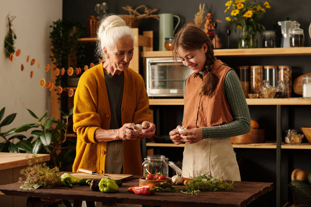 Senior woman explaining her granddaughter how to peel garlic for pickles while both standing by wooden table with jar containing marinade mix - Fotoğraf, Görsel