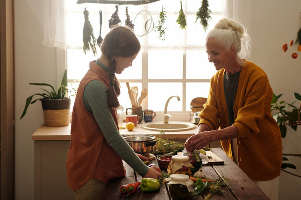 Vue latérale de la grand-mère et de la petite-fille heureuses faisant des cornichons ensemble tandis que la femme âgée met des légumes frais dans un pot - Photo, image
