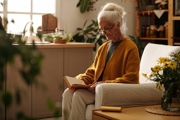 Komoly idős nő gyapjú kötött sárga kardigán olvasókönyv, miközben ül a nagy kényelmes karosszék a nappaliban a ház - Fotó, kép
