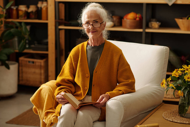 Glückliche Seniorin mit aufgeschlagenem Buch blickt in die Kamera, während sie im Sessel am kleinen Holztisch mit einem Strauß gelber Wildblumen sitzt - Foto, Bild
