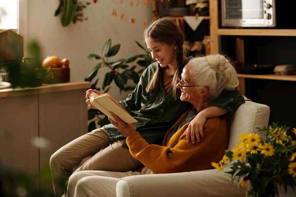 Вид сбоку на счастливую бабушку и внучку, читающих книгу рассказов или роман в свободное время, сидя в удобном кресле - Фото, изображение