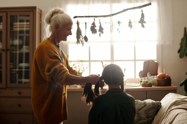 Side view of happy elderly woman with white hair plaiting braids to her granddaughter while standing in front of her against kitchen window - 写真・画像