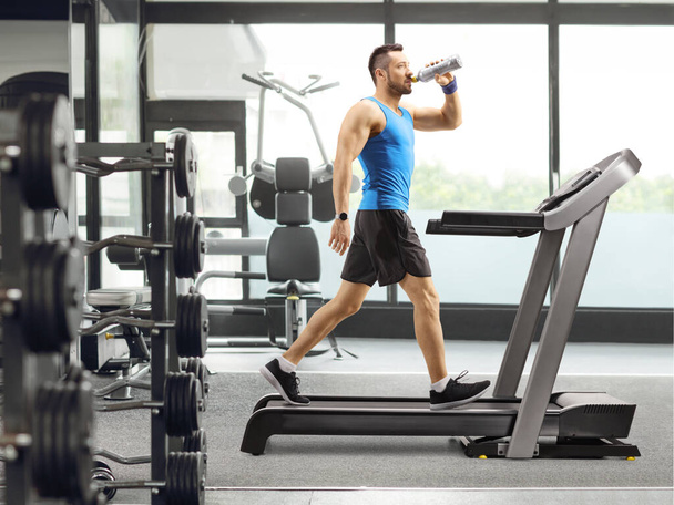 Full length profile shot of a young man running on a treadmill and drinking a sports energy drink at a gym - Foto, immagini