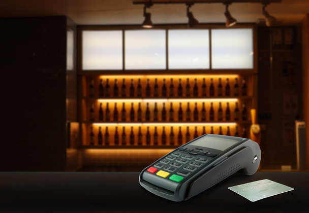 credit card reader pos machine and a credit card are on wooden table with background of beer bottle shelf in an empty nice bar in evening during cleaning process before opening to serve customers - Photo, Image