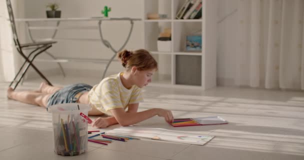 Une fille peint un tableau avec des crayons dans un album à la maison sur le sol. Enfant créativité et développement de la pensée et de la vision du monde. Images 4k de haute qualité - Séquence, vidéo