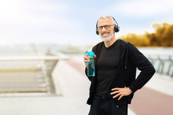 Positive handsome retired sportsman have outdoor workout at autumn park, happy senior man holding bottle, drinking water, hydration during exercising training concept, copy space - Photo, Image