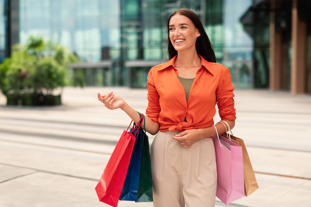 ショッピングを楽しんでいる陽気で美しい千年紀ヨーロッパの女性は,買い物で多くの紙パッケージやカラフルな買い物バッグを持ち,外を見ながら都市部を歩いています. シーズンセール - 写真・画像
