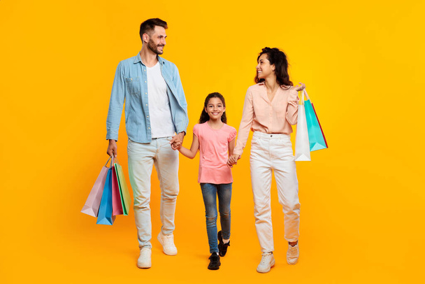 Οικογενειακή ιδέα αγορών. Χαρούμενοι Καυκάσιοι γονείς και η κόρη μεταφέρουν τσάντες, κρατώντας τα χέρια και περπατώντας μαζί σε κίτρινο φόντο, σε όλο το μήκος - Φωτογραφία, εικόνα