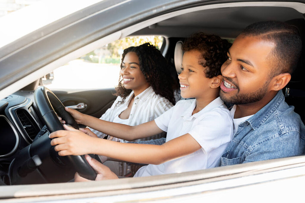 Glückliche afrikanisch-amerikanische Eltern Vater und Mutter genießen nagelneues Auto mit ihrem kleinen Sohn. Aufgeregt sitzt ein schwarzer Teenager auf Papas Schoß, fährt Auto und lächelt - Foto, Bild