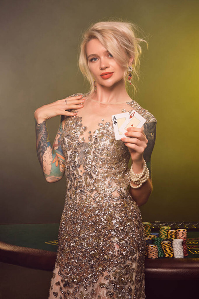 Κοντινό πλάνο μιας όμορφης γυναίκας με ξανθά μαλλιά, τατουάζ στα χέρια και τέλειο μακιγιάζ, ντυμένη με ασημί γυαλιστερό φόρεμα. Στέκεται πάνω σε ένα τραπέζι τζόγου, με δύο τραπουλόχαρτα στα χέρια της και - Φωτογραφία, εικόνα