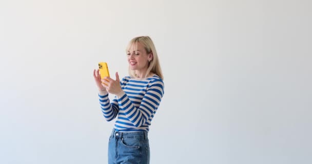 Cheerful and radiant Caucasian woman is seen on a clean white backdrop, holding a phone and bursting into laughter. Her infectious smile and joyful expression show her genuine amusement. - Footage, Video