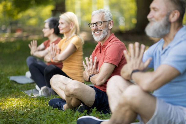 Portret przystojnego starszego mężczyzny uczęszczającego na zajęcia jogi grupowej na świeżym powietrzu, szczęśliwy dojrzały dżentelmen praktykujący medytację z przyjaciółmi w parku, siedzący w pozycji lotosu i uśmiechnięty przed kamerą - Zdjęcie, obraz