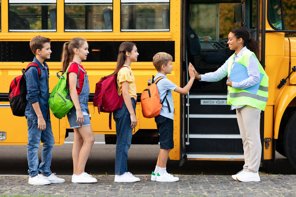 Nauczyciel w mundurze daje przybijając piątkę do dzieci wchodzących do autobusu szkolnego, przyjazny czarny asystent pani powitanie dzieci w pobliżu autobusu szkolnego, szczęśliwych chłopców i dziewcząt stojących w rzędzie, czeka skręcić razem - Zdjęcie, obraz