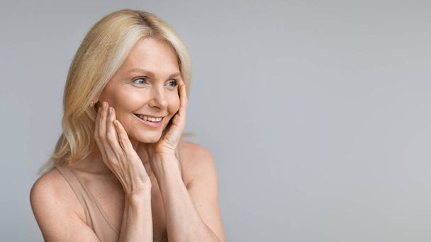 Beautiful mature woman looking aside at copy space and touching cheeks, smiling attractive aged lady enjoying skincare treatments, standing over grey studio background, panorama - Photo, Image