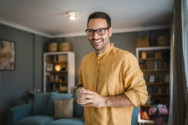 Один молодой кавказский мужчина пьет кофе, стоя дома в рубашке по утрам реальные люди копируют пространство - Фото, изображение