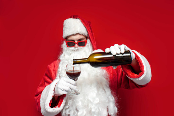 drunk santa claus in hat and festive glasses drinks wine from bottle on red background, man in santa costume drinks alcohol for christmas - Photo, Image