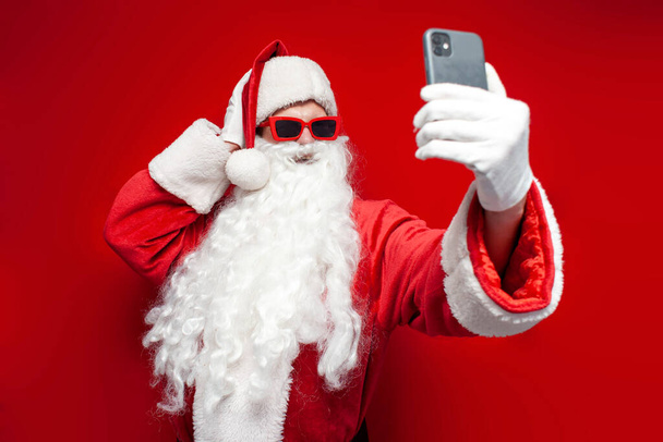 Babbo Natale in cappello e occhiali festivi utilizza smartphone e prende selfie su sfondo rosso, l'uomo in costume di Babbo Natale parla tramite link video online - Foto, immagini