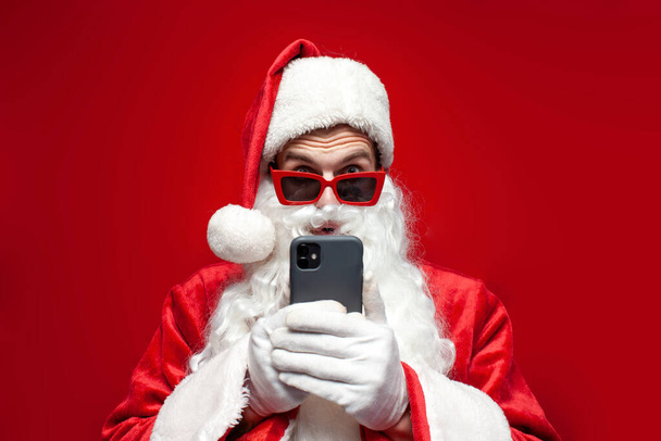 shocked santa claus in hat and festive glasses uses smartphone and is surprised on red background, amazed man in santa costume is typing online on the phone at christmas - Photo, Image