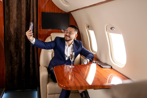 Молодий азійський бізнесмен сидить на приватному розкішному літаку і спілкується за допомогою смартфона відео дзвінка, фінансового менеджера в костюмі і окулярах, розмовляючи по телефонній відео. - Фото, зображення