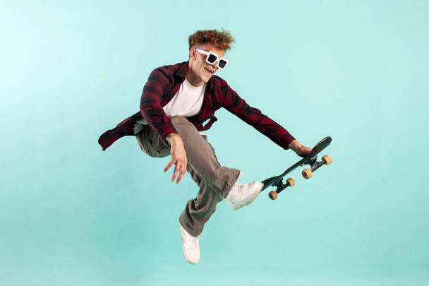 young crazy guy rides skateboard and jumps on blue isolated background, hipster in sunglasses flies with skateboard in the air and does extreme trick - Photo, Image