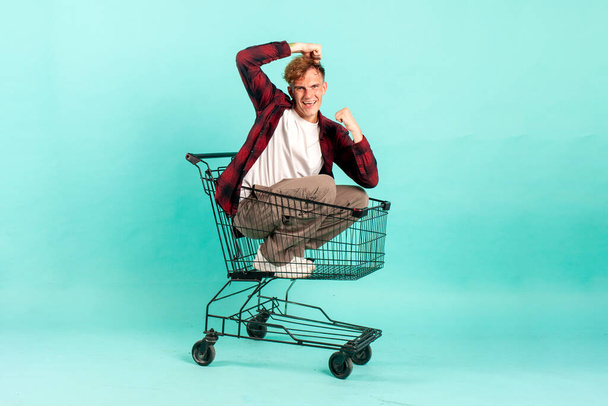 young cheerful guy winner sits in shopping cart and rejoices in victory on blue background, student buyer goes to the supermarket in empty cart - Photo, Image