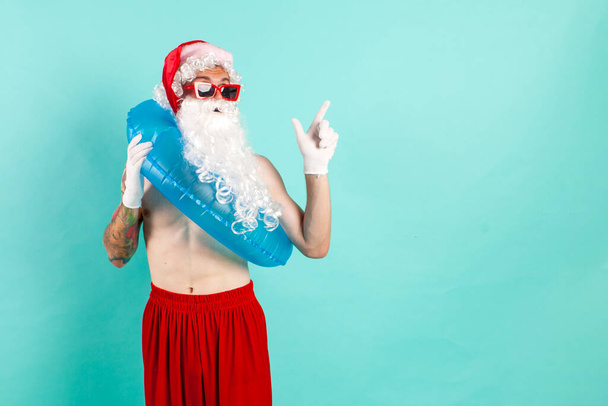 Санта-Клаус в костюме с надувным кольцом для плавания в солнцезащитных очках лежит на пляже и указывает на пространство для копирования на голубом изолированном фоне, Санта-туристические аттракционы на отдыхе - Фото, изображение