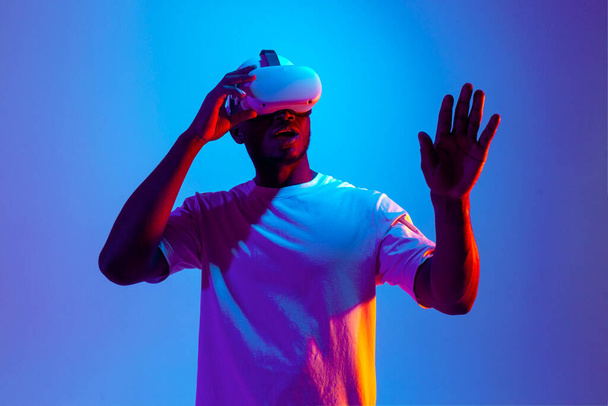 african american male gamer in virtual reality glasses touches his hands in neon lighting, the guy uses modern vr gadget and chooses and points his finger at copy space - Photo, Image