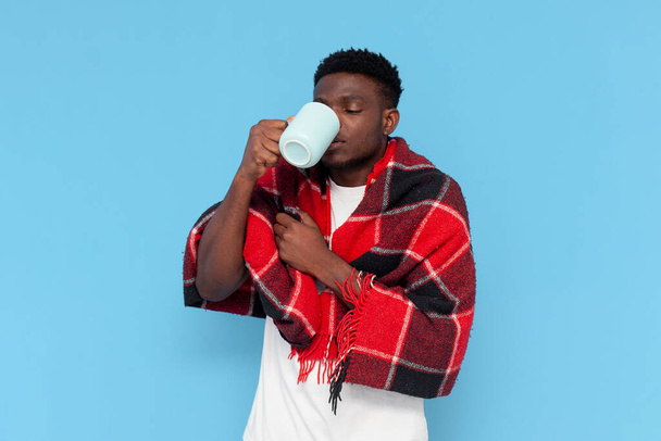 Kranker junger Mann aus Afrika trinkt Medizin und leidet an Fieber und hüllt sich in eine warme Decke auf blauem isolierten Hintergrund, Mann mit Getränk zeigt Erkältungs- und Grippesymptome - Foto, Bild