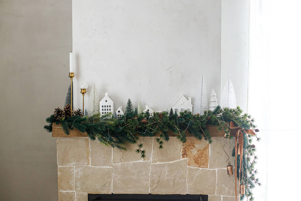 Elegante chimenea de Navidad decorada. Modernos árboles de Navidad, casas, conos de pino y ramas de abeto en la chimenea mantel con campanas de latón y cinta. Navidad escandinava decoración de la sala de estar - Foto, Imagen