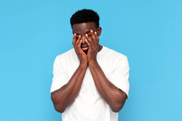 κουρασμένος Αφροαμερικάνος με λευκό μπλουζάκι σε κατάθλιψη καλύπτει το πρόσωπό του με τα χέρια του σε μπλε απομονωμένο φόντο, νυσταγμένος αδιάφορος άνθρωπος βαριέται και δυστυχισμένος - Φωτογραφία, εικόνα